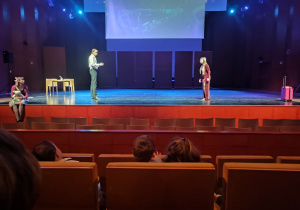 Fragment przedstawienia. Na scenie śpiewają dwie osoby jedna siedzi na podłodze i słucha.