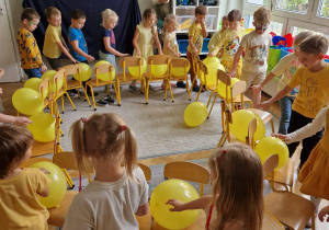 Dzieci w sali chodzą na około ustawionych krzesełek trzymając żółte balony
