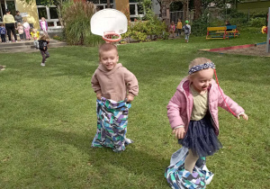 Dwoje dzieci skacze w workach podczas przedszkolnych zawodów na trawie.