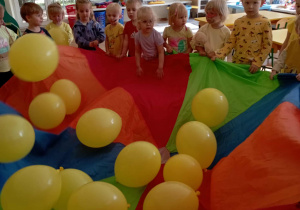Dzieci trzymają kolorową chustę animacyjną z żółtymi balonami