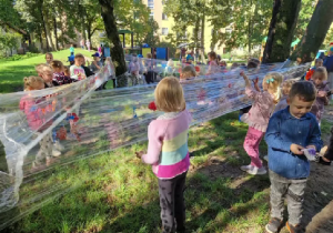 Dzieci wspólnie malują farbami na rozwieszonej między drzewami foli.