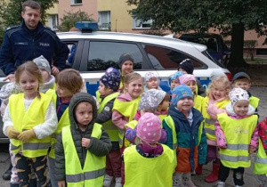 Dzieci w kazmizelkach odblaskowych stoją przed radiowozem razem z Panem Policjantem
