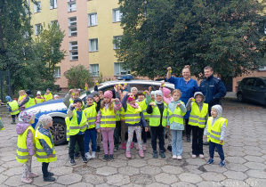 Dzieci z grupy Sowy stoją przed radiowozem razem z Wychowawcą i Panem Policjantem pokazując odblaski
