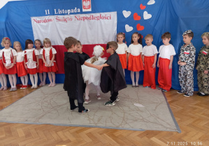 Chłopcy w czarnych pelerynach trzymająBiałego orła, w tle dzieci z grupy Sowy