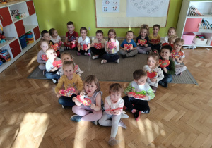 Dzieci z grupy Żabki siedzę na dywanie w kształcie serca trzymając w dłoniach swoje czerwone serduszka