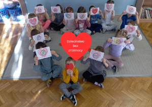 dzieci siedzą na dywanie w kształcie serca pokazując swoje prace