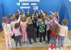 Dzieci stoją przodzem do dekoracji wskazując ręką napis Unicef