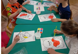 Dzieci przy stoliku malują farbami obrazek misia