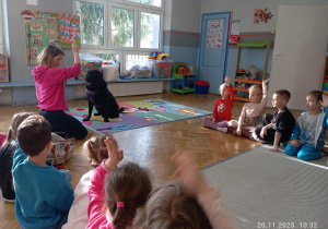 Dzieci siedzą na dywanie, opiekunka wydaje komendy psu.