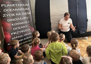 Dzieci siedzą na dywanie słuchają prowadzącej, która opowiada o planetacho planetach