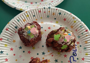 Na papierowych talerzykach leża papierowe pączki udekorowane lukrem zrobionym z białej farby oraz posypką kolorowego papieru.