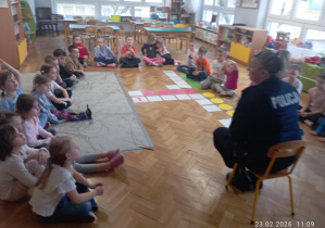 dzieci siedzą na dywanie i słuchają Pani Policjantki, która tłumaczy jak być bezpiecznym np; przechodząc przez jezdnię.