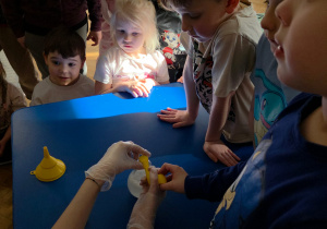 Dzieci obserwują doświadczenie z wodą i balonem wykonywane przez prowadzącą przy stoliku.