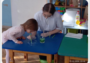 Dziewczynka obserwuje bańkę, a prowadząca pokazuje eksperyment z użyciem słomki i płynu.