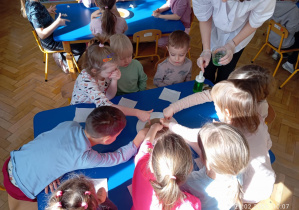 Grupa dzieci siedzi przy stoliku i wspólnie uczestniczy w doświadczeniu z wodą pod okiem prowadzącej.