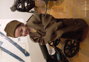 Dziewczynka w wojskowym płaszczu i czapce uśmiecha się, stojąc obok eksponatu.