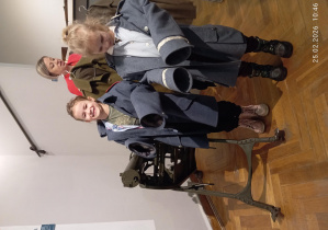 Dziewczynki w dużych mundurach wojskowych stoi obok eksponatu pod opieką prowadzącej.