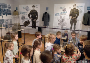 Grupa dzieci ogląda eksponaty wojskowe w gablotach podczas zwiedzania muzeum.