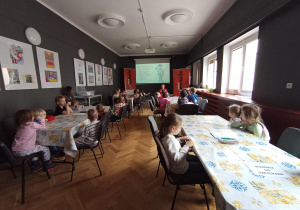 Dzieci siedzą przy stołach i uczestniczą w warsztatach edukacyjnych w sali muzealnej.