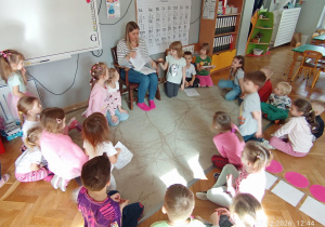 Dzieci siedzą w kole na dywanie a pani Monika rozdaje im kolorowanki z postaciami z bajek Hansa Christiana Andersena