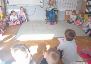 Dzieci siedzą w kole na dywanie i słuchają czytanej bajki.
