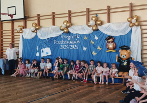 Dzieci siedzą przed dekoracją przygotowaną na zakończenie roku przedszkolnego.